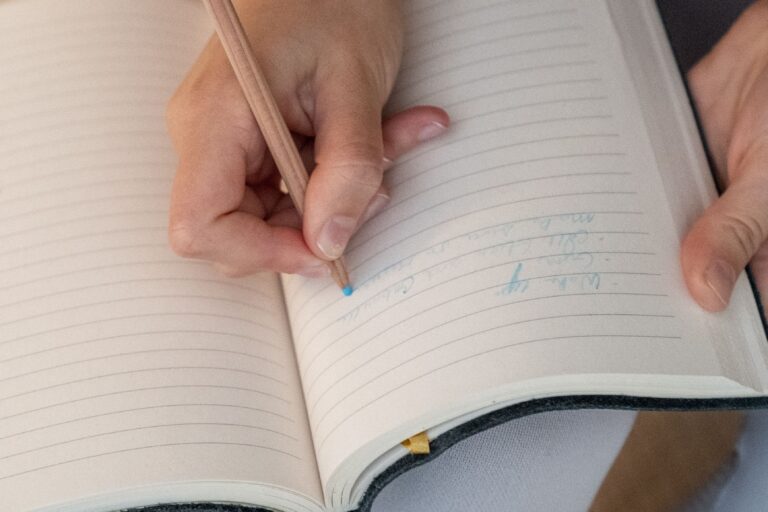 A hand writes in an open lined notebook with a blue pencil, showcasing a close-up of the writing process.